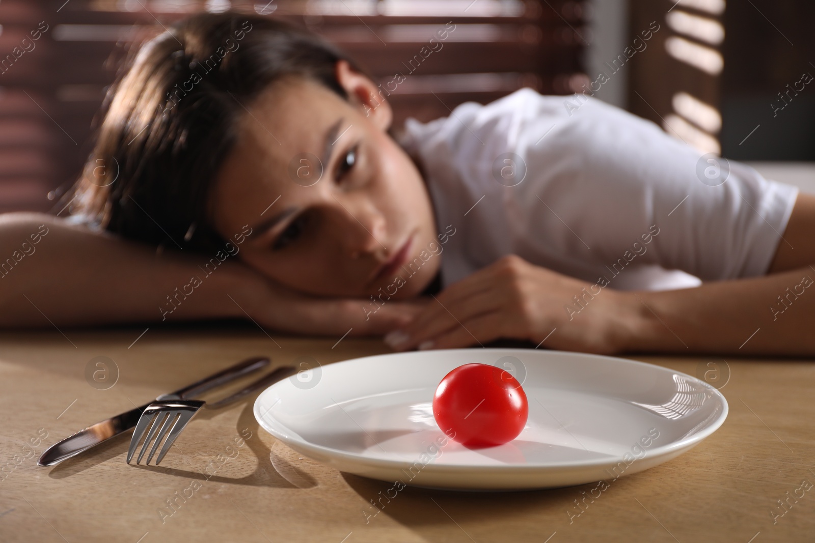 Photo of Eating disorder. Sad woman at wooden table with cutlery, tomato and plate indoors