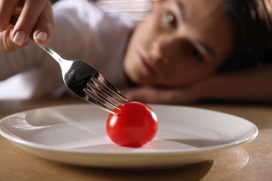 Eating disorder. Sad woman holding fork with tomato at wooden table indoors, selective focus
