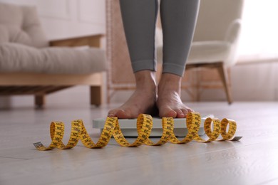 Photo of Eating disorder. Woman standing on floor scale and measuring tape indoors, closeup