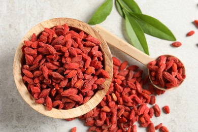 Photo of Dried goji berries, bowl, spoon and green leaves on light textured table, flat lay