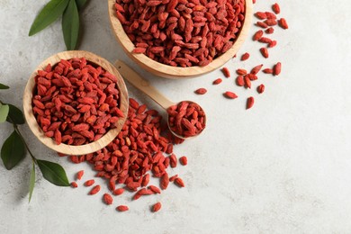 Dried goji berries, bowl, spoon and green leaves on light textured table, flat lay. Space for text