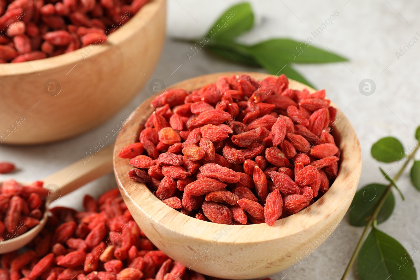 Photo of Dried goji berries, bowl and green leaves on light textured table, closeup