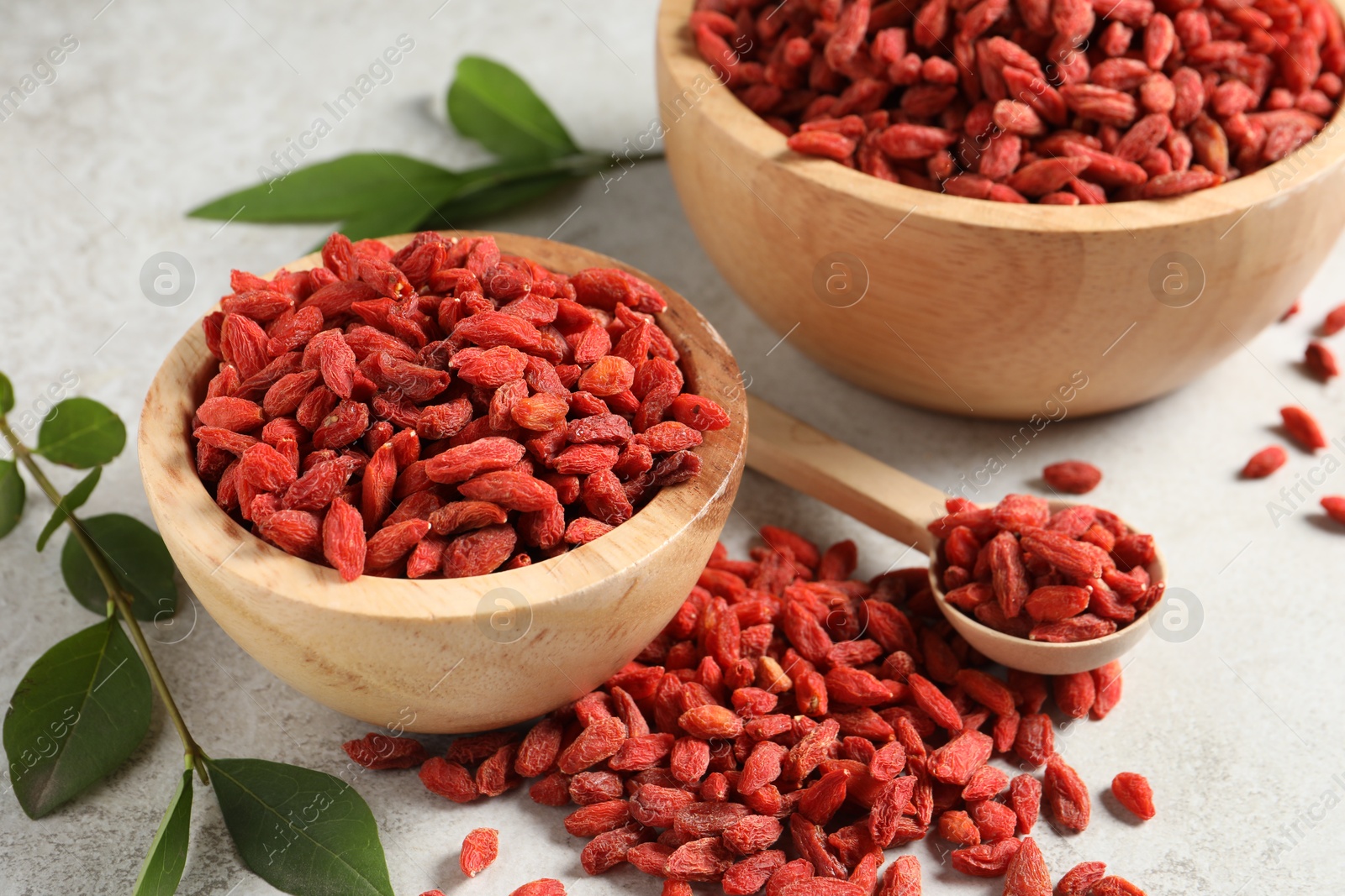 Photo of Dried goji berries, bowl and green leaves on light textured table, closeup