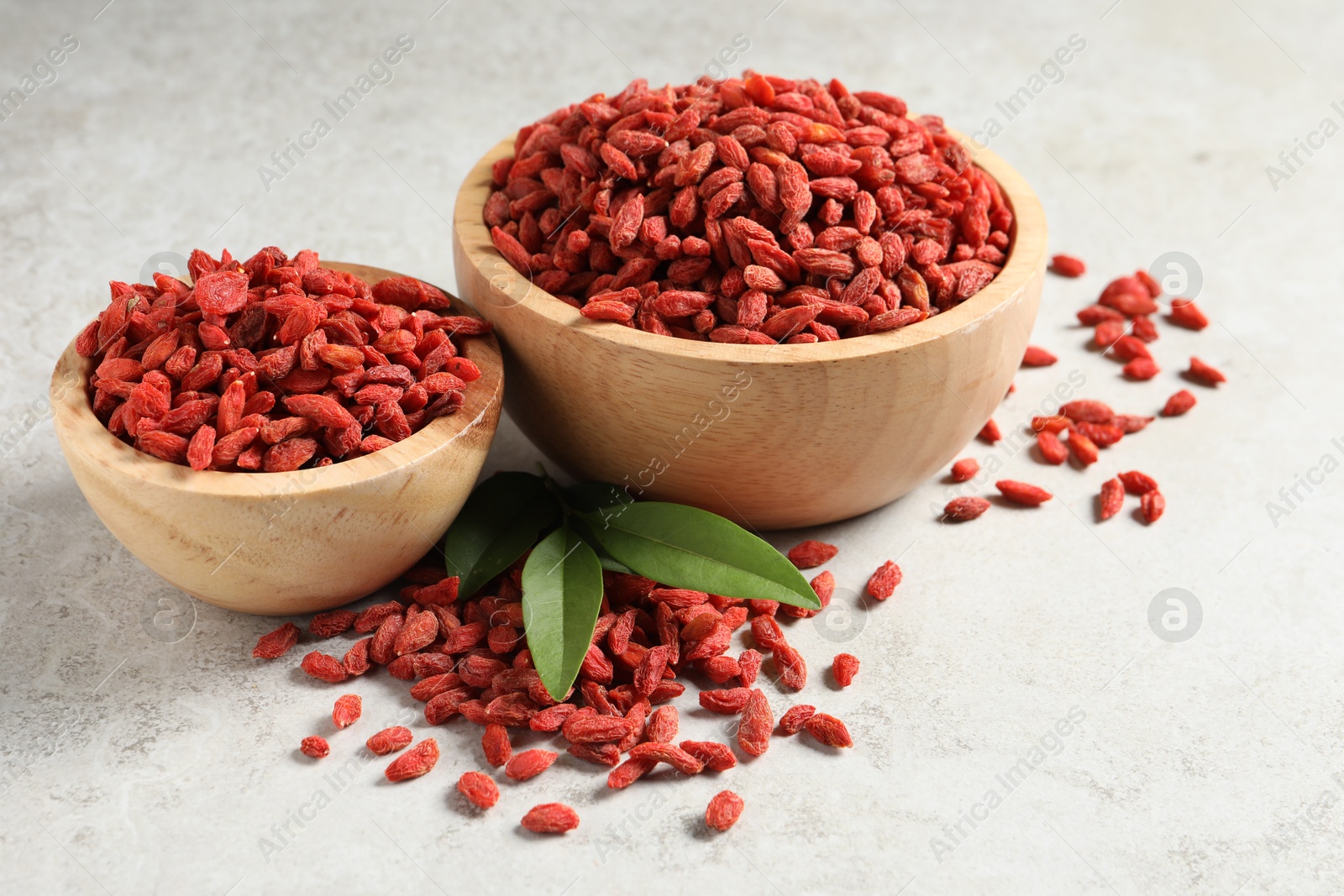 Photo of Dried goji berries, bowls and green leaves on light textured table, closeup