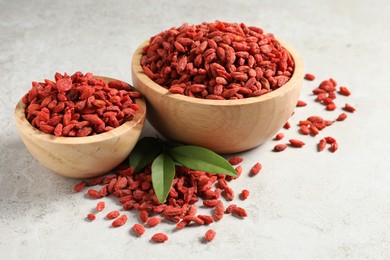 Dried goji berries, bowls and green leaves on light textured table, closeup