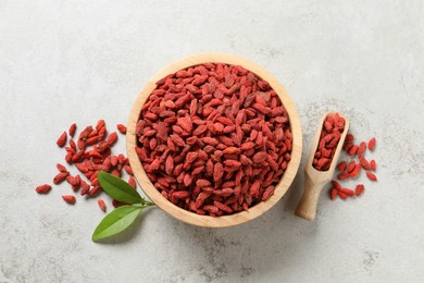 Dried goji berries in bowl, scoop and green leaves on light textured table, flat lay