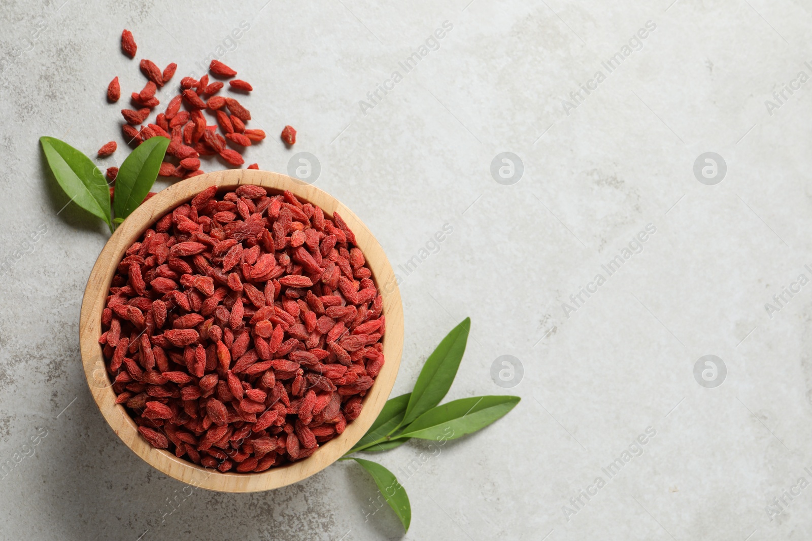 Photo of Dried goji berries in bowl and green leaves on light textured table, flat lay. Space for text