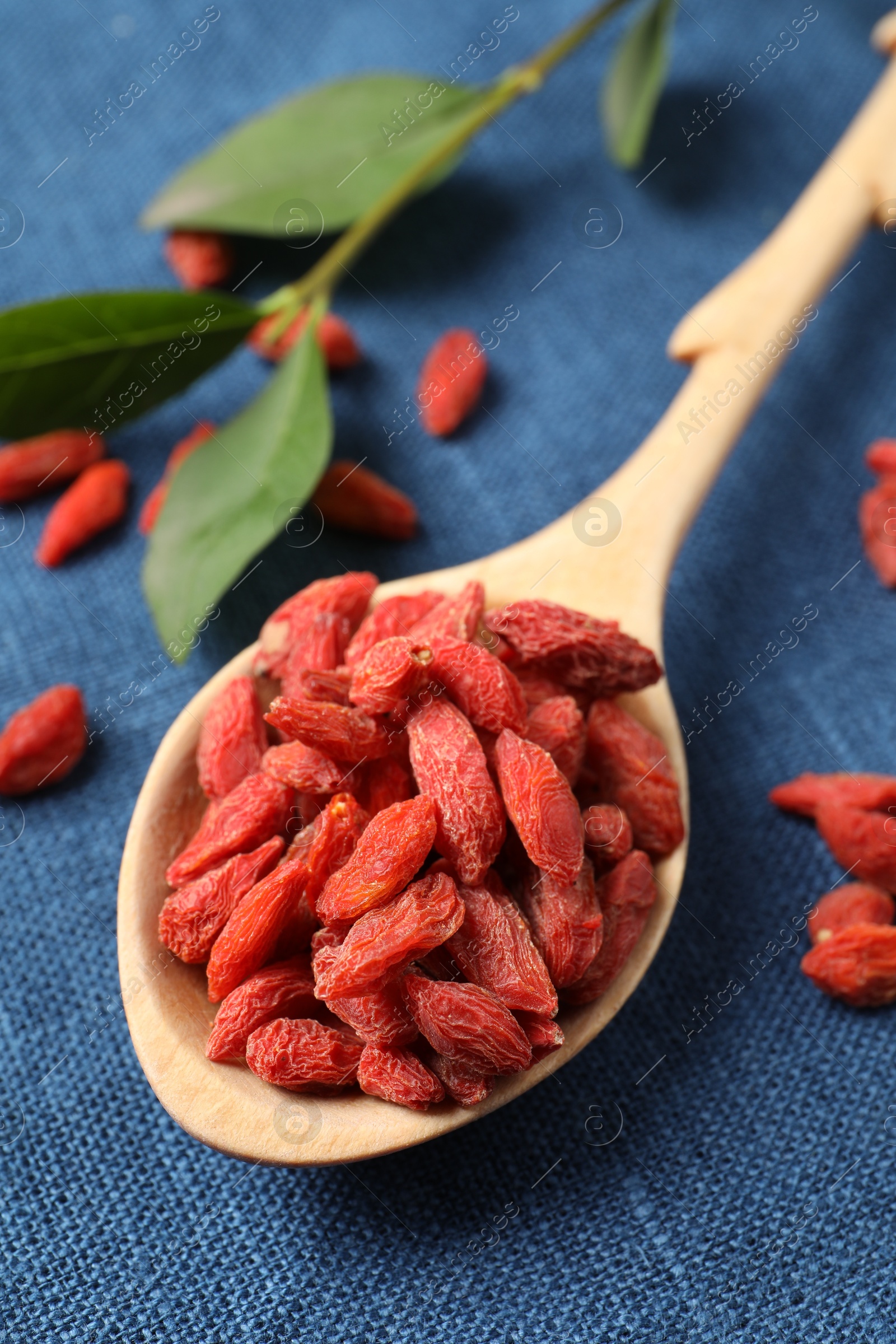 Photo of Spoon with dried goji berries and green leaves on blue cloth, closeup