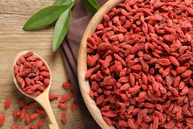 Dried goji berries in bowl and spoon on wooden table, flat lay