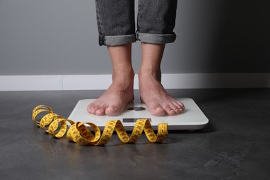 Photo of Eating disorder. Woman standing on floor scale and measuring tape indoors, closeup