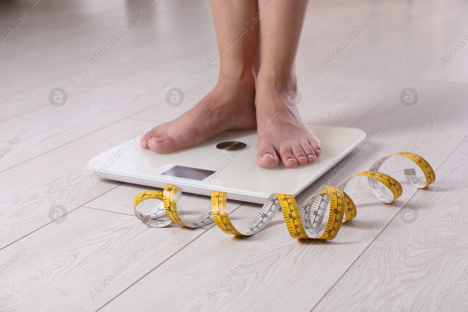 Photo of Eating disorder. Woman standing on floor scale and measuring tape indoors, closeup