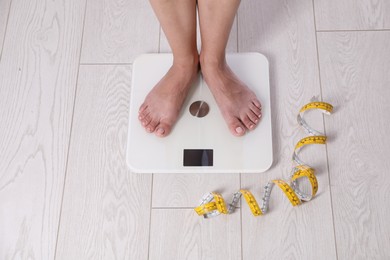 Photo of Eating disorder. Woman standing on floor scale and measuring tape indoors, above view
