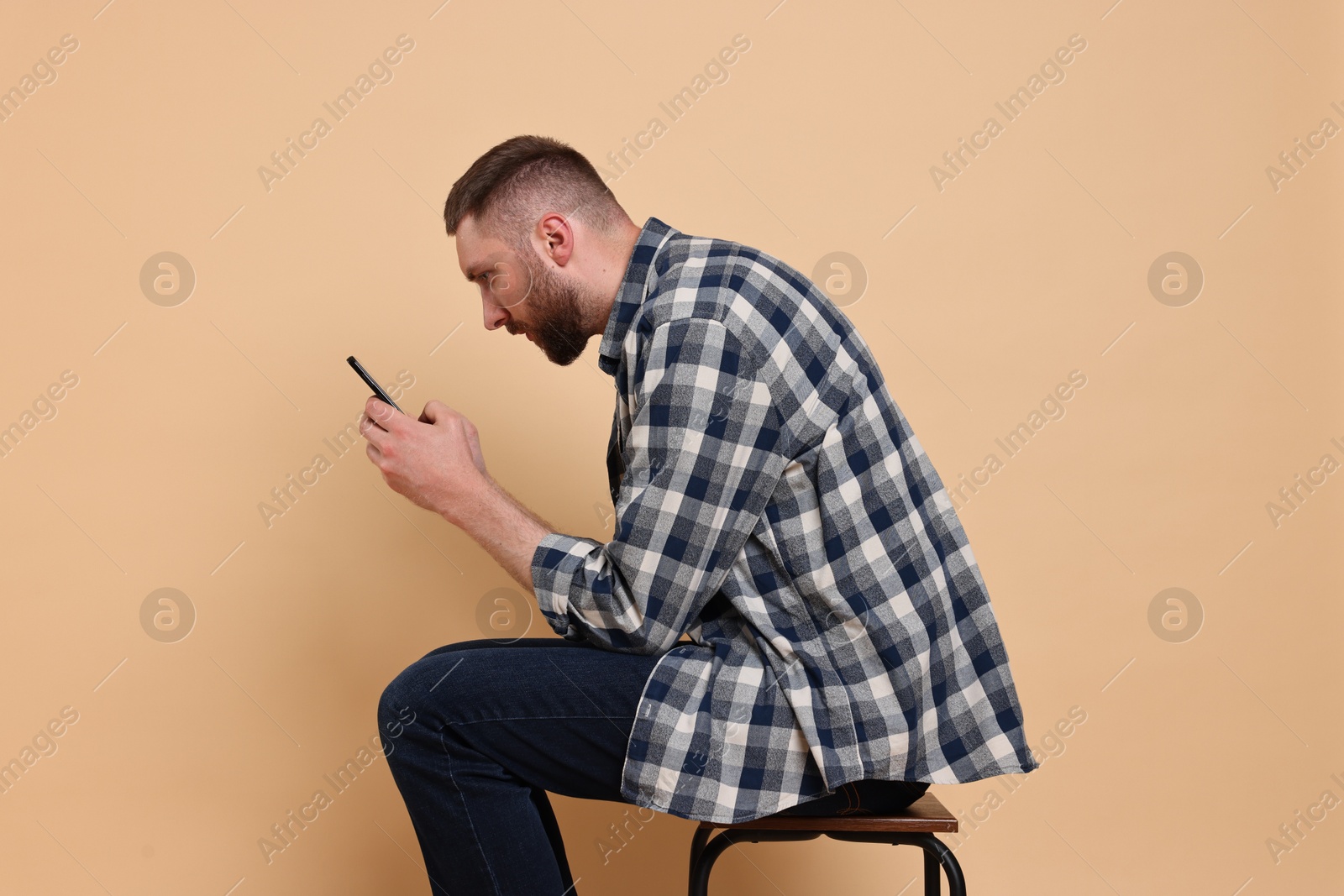 Photo of Man with poor posture sitting on chair and using smartphone against pale orange background