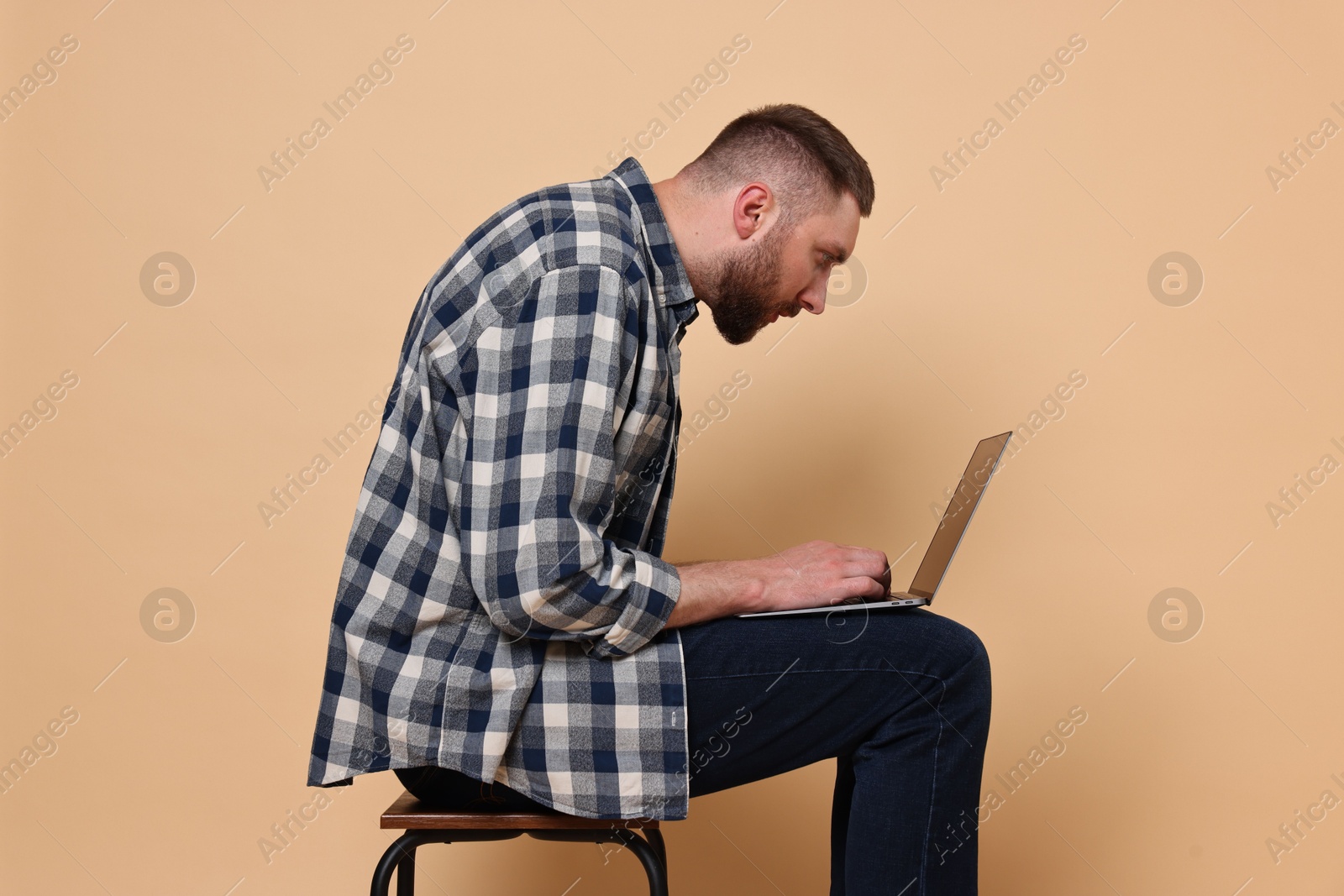 Photo of Man with poor posture sitting on chair and using laptop against pale orange background