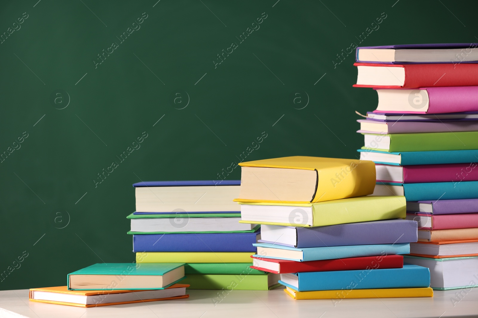 Photo of Stacks of many colorful books on white table near chalkboard in classroom. Space for text