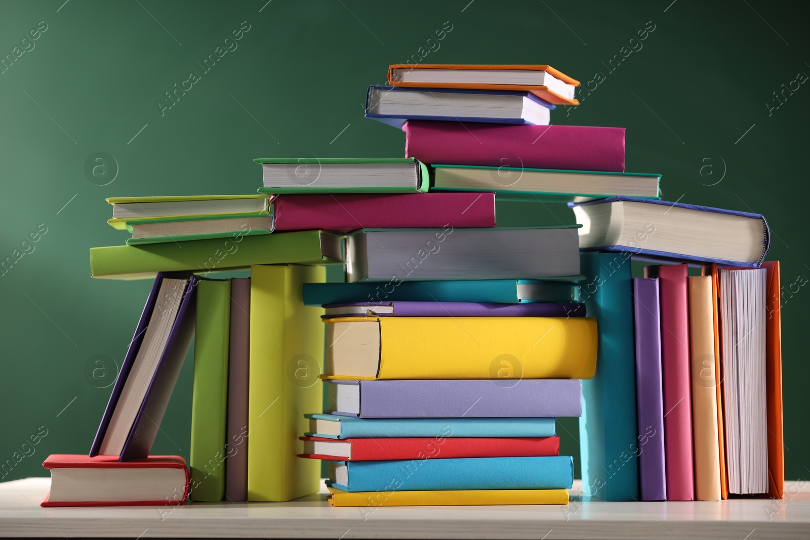 Photo of Stacks of many colorful books on white wooden table near chalkboard in classroom