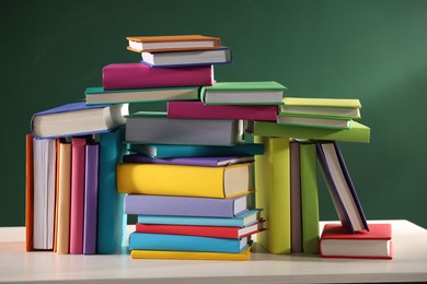 Photo of Stacks of many colorful books on white wooden table near chalkboard in classroom