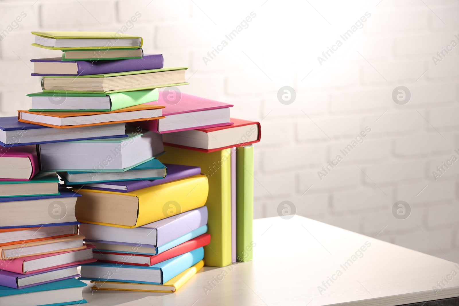 Photo of Stacks of many colorful books on white wooden table against brick wall. Space for text