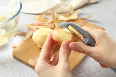 Woman peeling fresh potato with peeler at light grey table, closeup