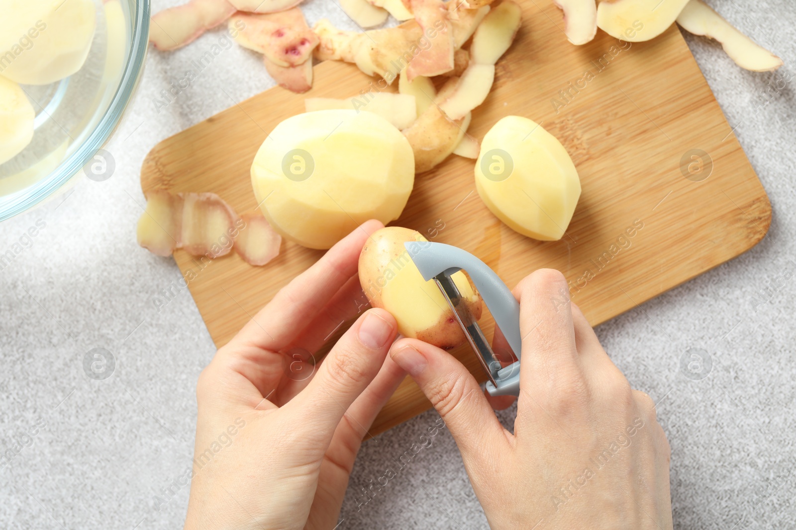 Photo of Woman peeling fresh potato with peeler at light grey table, top view