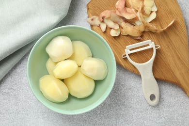 Photo of Fresh raw potatoes in bowl, peeler and peels on light grey table, top view