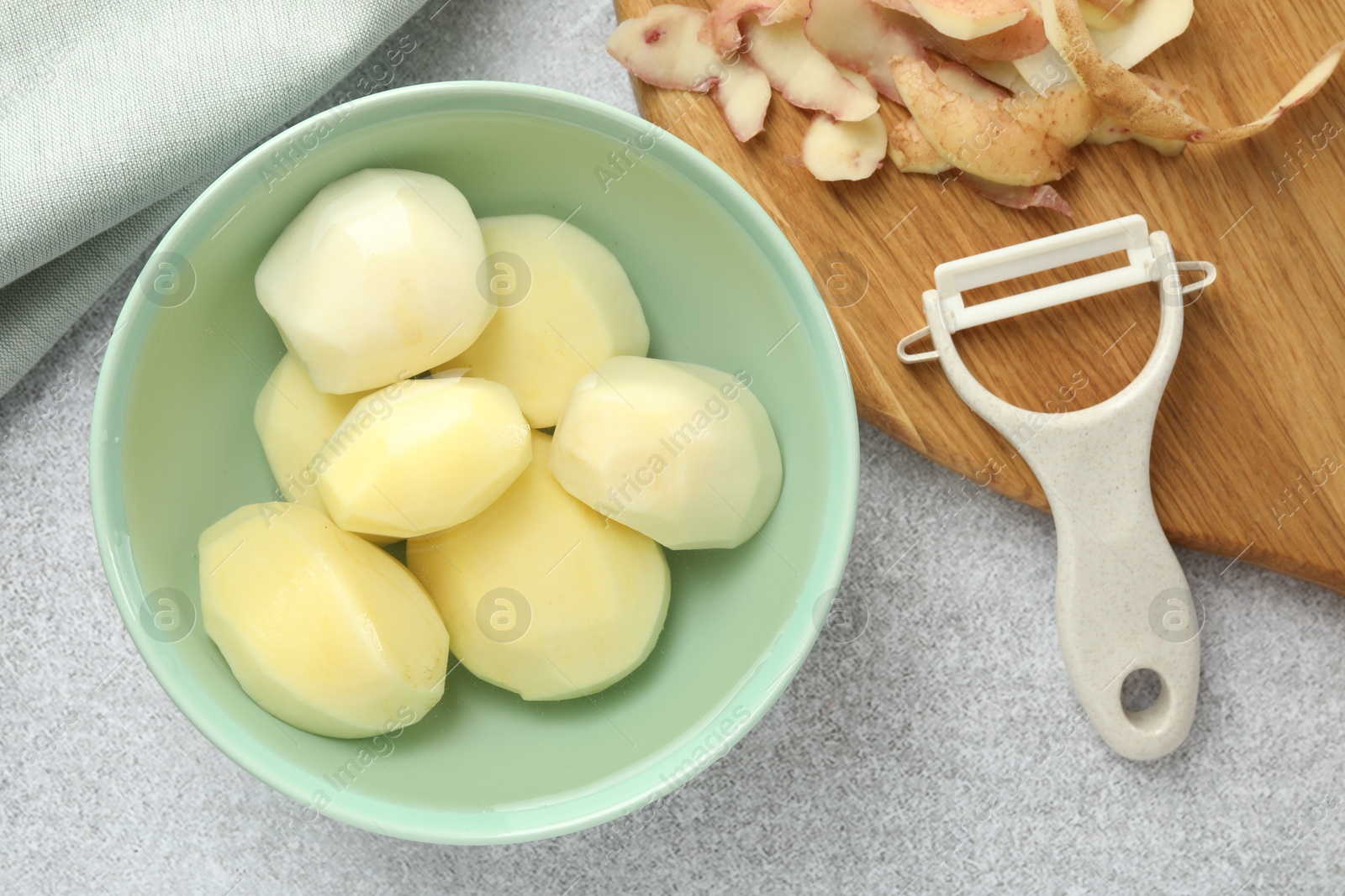 Photo of Fresh raw potatoes in bowl, peeler and peels on light grey table, top view