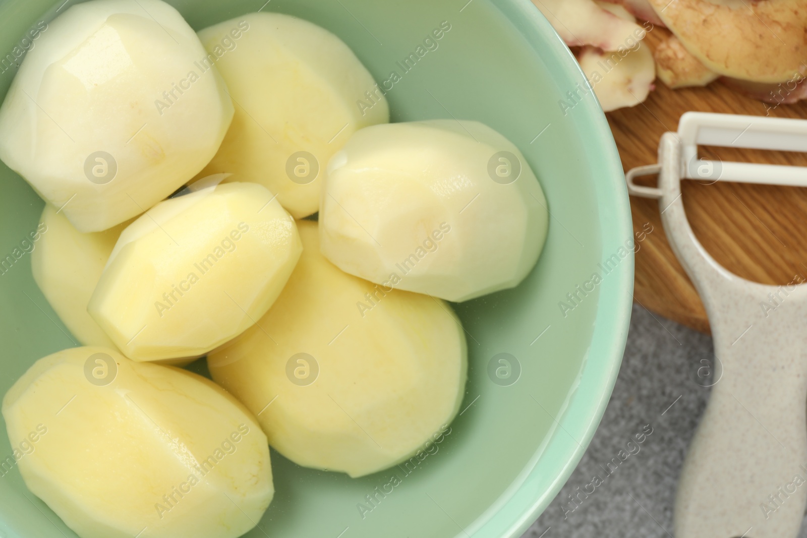 Photo of Fresh raw potatoes in bowl, peeler and peels on light grey table, top view