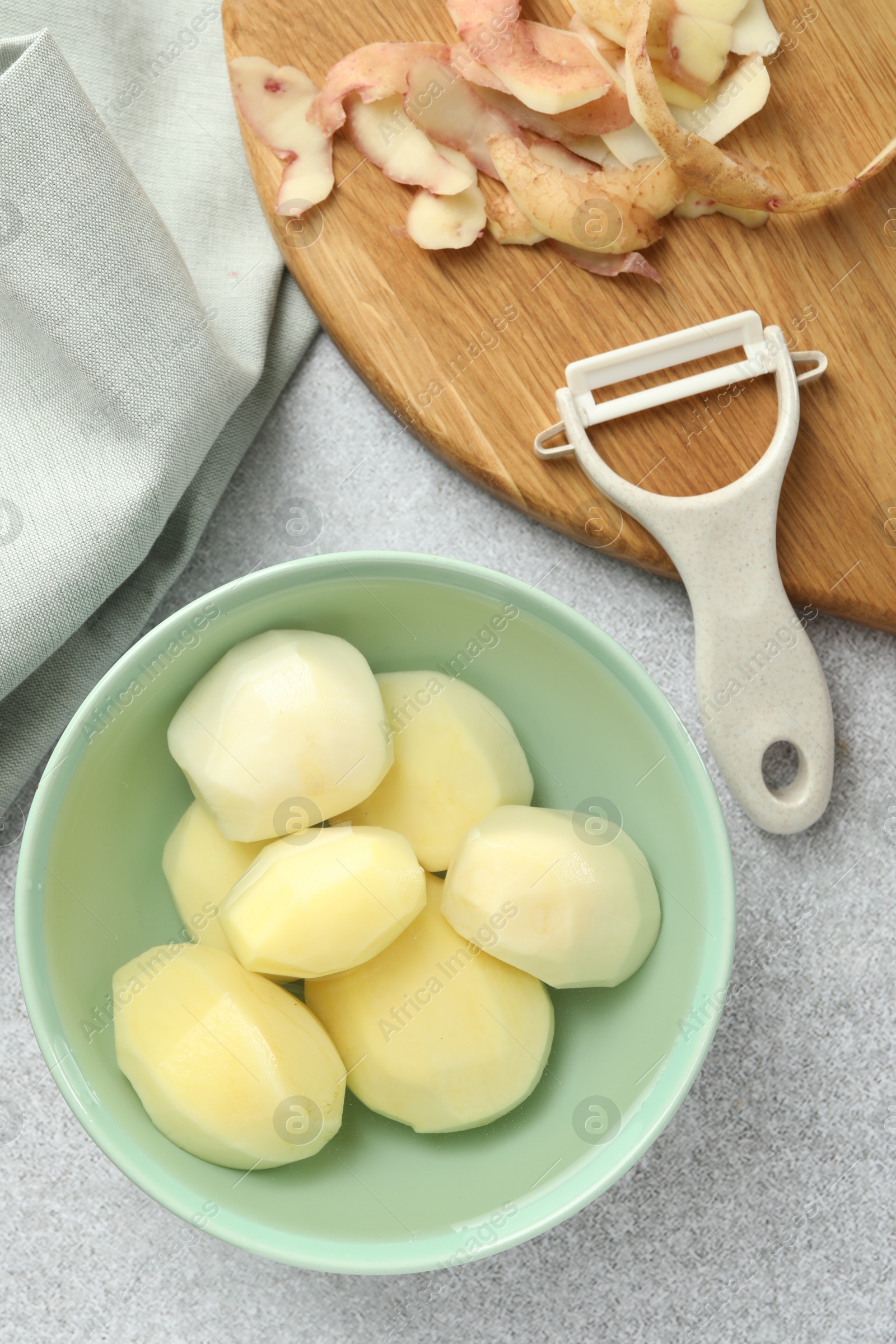 Photo of Fresh raw potatoes in bowl, peeler and peels on light grey table, top view