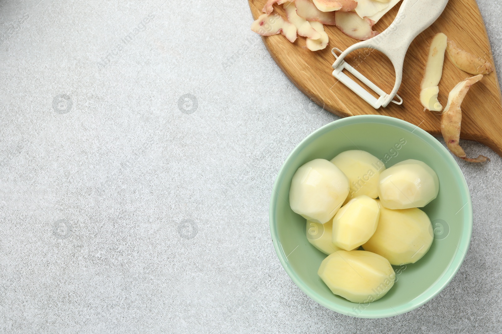 Photo of Fresh raw potatoes in bowl, peeler and peels on light grey table, top view. Space for text