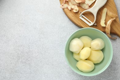 Fresh raw potatoes in bowl, peeler and peels on light grey table, top view. Space for text