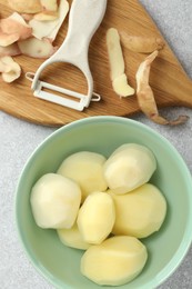 Photo of Fresh raw potatoes in bowl, peeler and peels on light grey table, top view