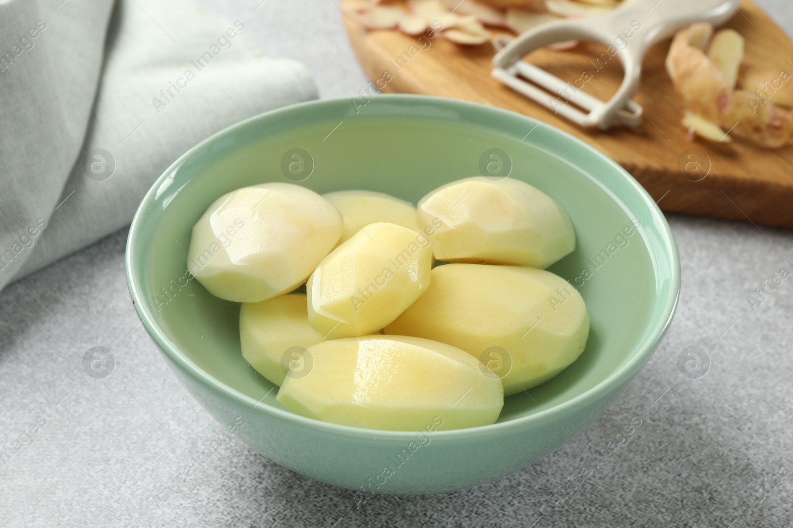 Photo of Fresh raw potatoes in bowl, peeler and peels on light grey table