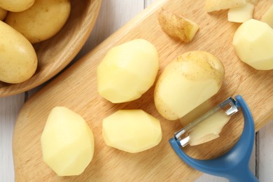 Photo of Fresh raw potatoes, peels and peeler on white wooden table, top view