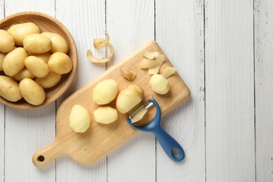 Photo of Fresh raw potatoes, peels and peeler on white wooden table, top view. Space for text