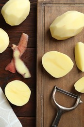 Fresh raw potatoes, peels and peeler on wooden table, top view