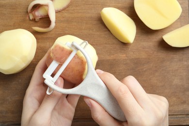 Photo of Woman peeling fresh potato with peeler at wooden table, top view