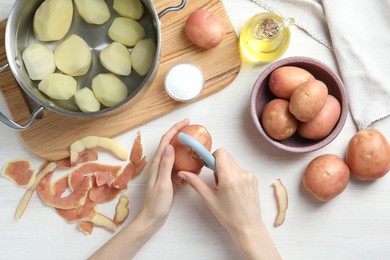 Photo of Woman peeling fresh potato with peeler at white wooden table, top view