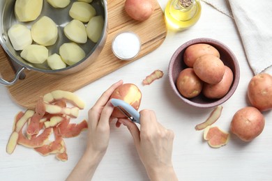 Photo of Woman peeling fresh potato with peeler at white wooden table, top view