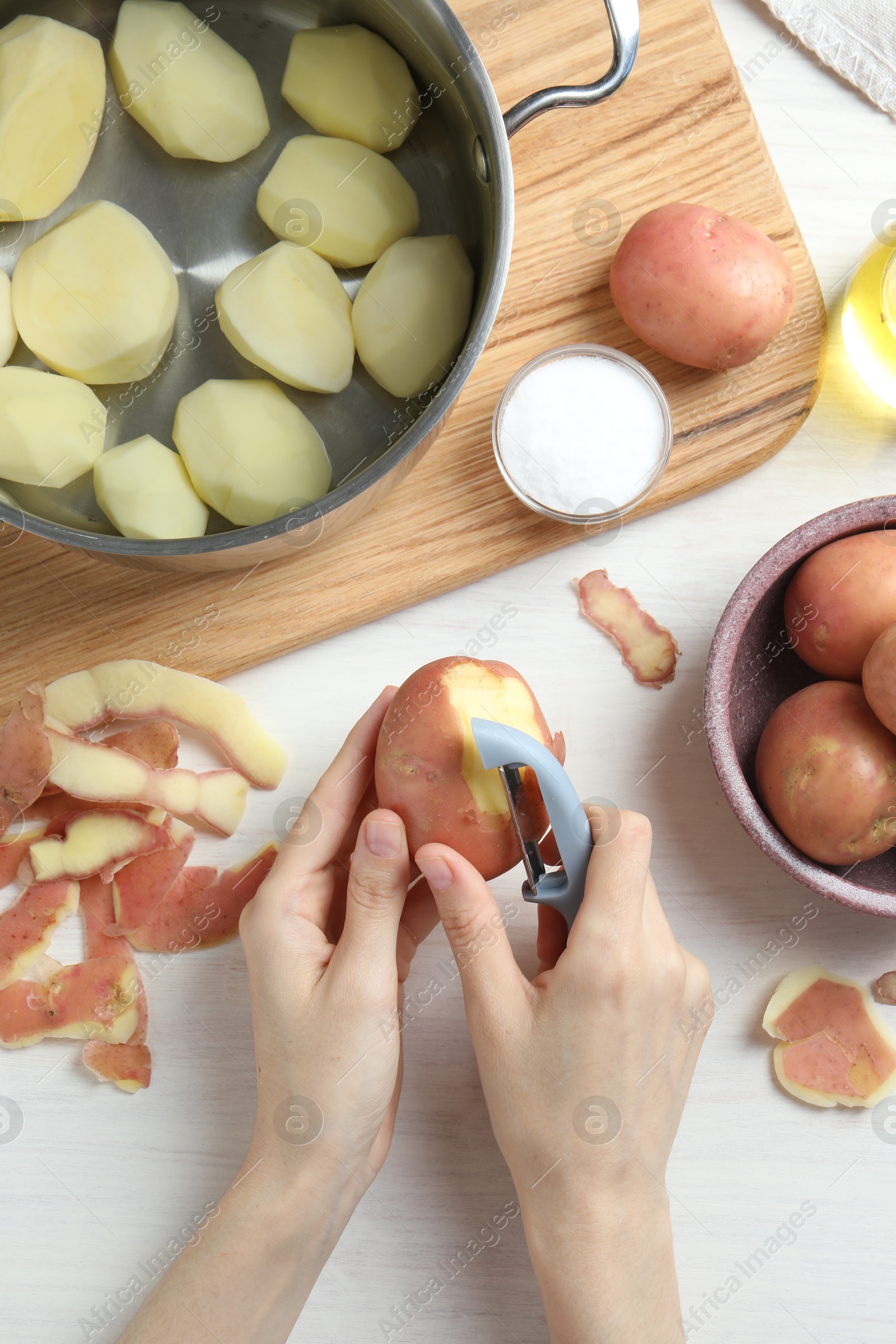 Photo of Woman peeling fresh potato with peeler at white wooden table, top view