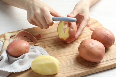 Woman peeling fresh potato with peeler at white wooden table, closeup