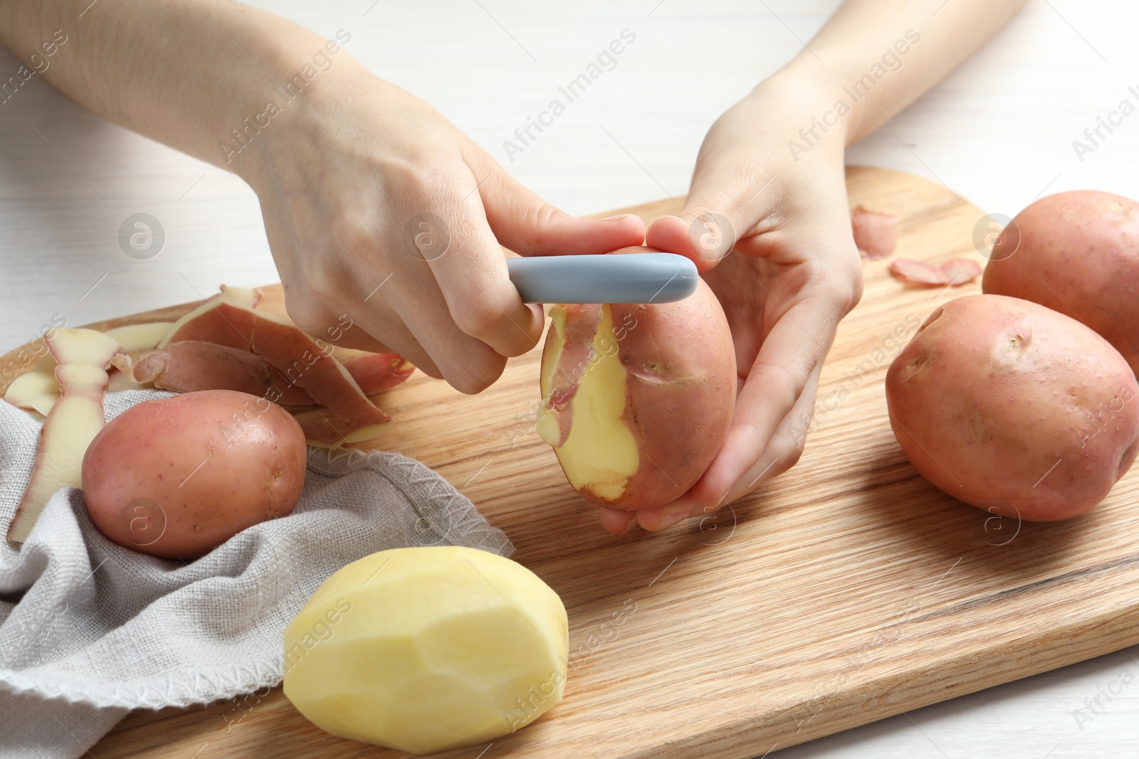 Photo of Woman peeling fresh potato with peeler at white wooden table, closeup