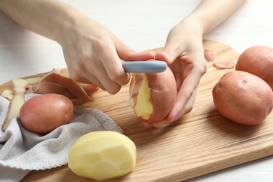 Woman peeling fresh potato with peeler at white wooden table, closeup