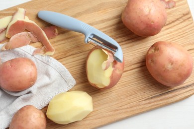 Fresh raw potatoes, peels and peeler on white table, top view