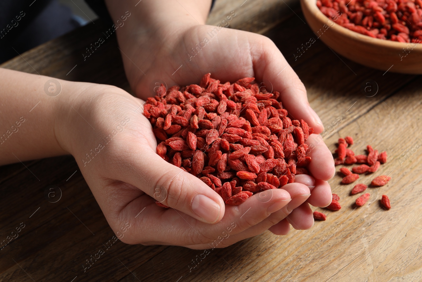 Photo of Woman holding pile of dried goji berries at wooden table, closeup