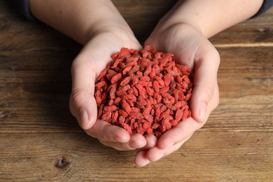 Photo of Woman holding pile of dried goji berries at wooden table, closeup