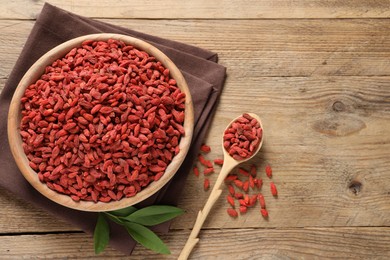 Photo of Dried goji berries in bowl and spoon on wooden table, flat lay. Space for text