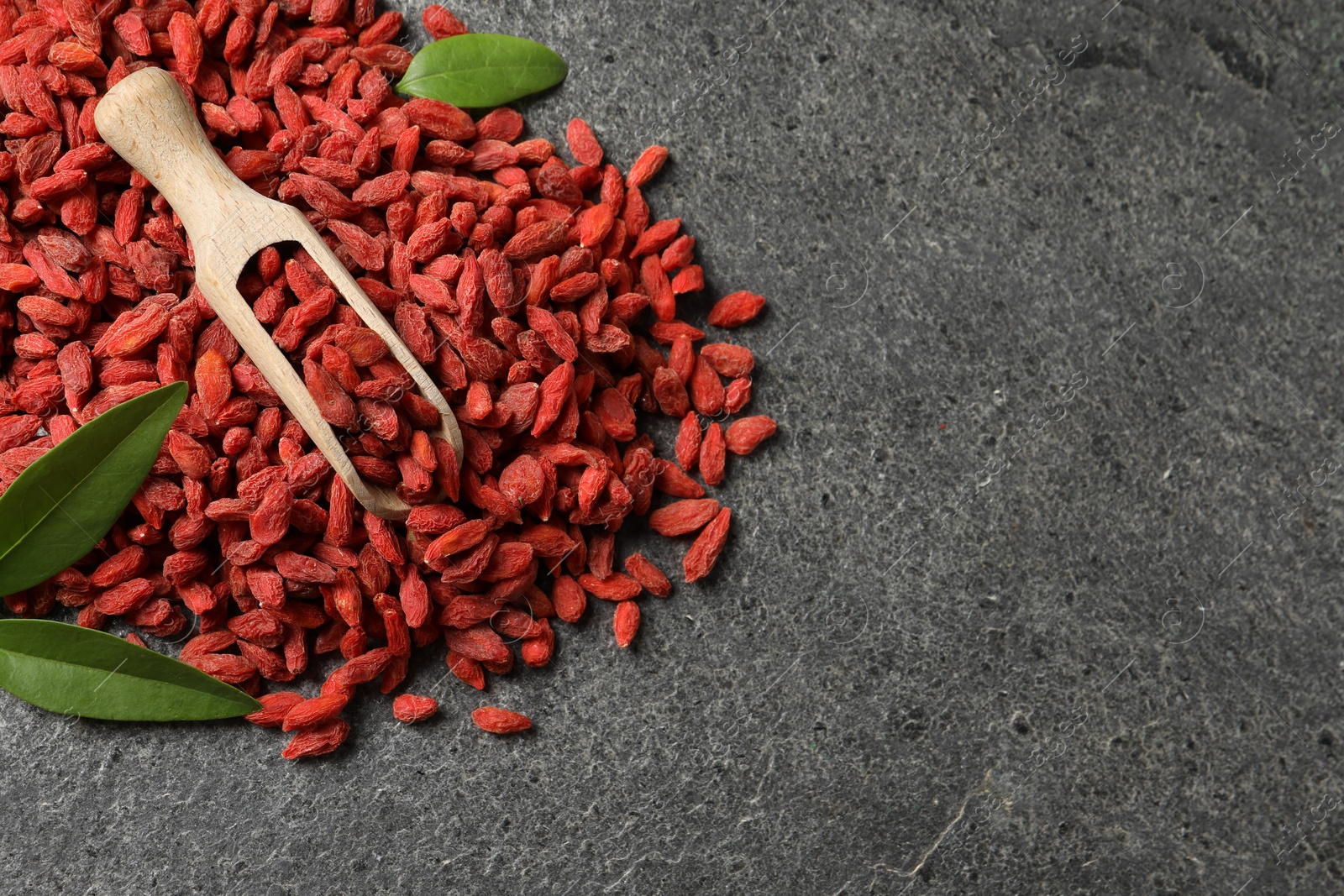 Photo of Dried goji berries, leaves and scoop on grey textured table, flat lay. Space for text