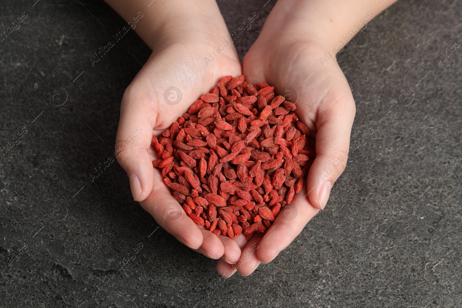 Photo of Woman holding pile of dried goji berries on grey textured table, top view