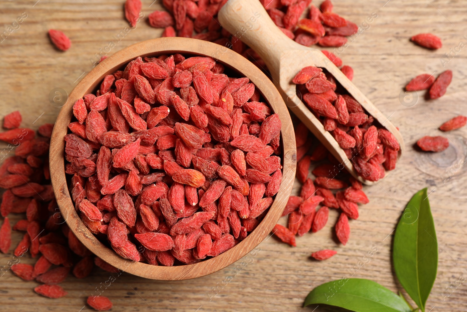 Photo of Dried goji berries, leaves, bowl and scoop on wooden table, flat lay
