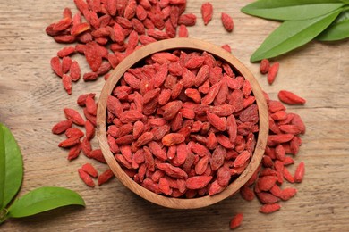 Dried goji berries, leaves and bowl on wooden table, flat lay